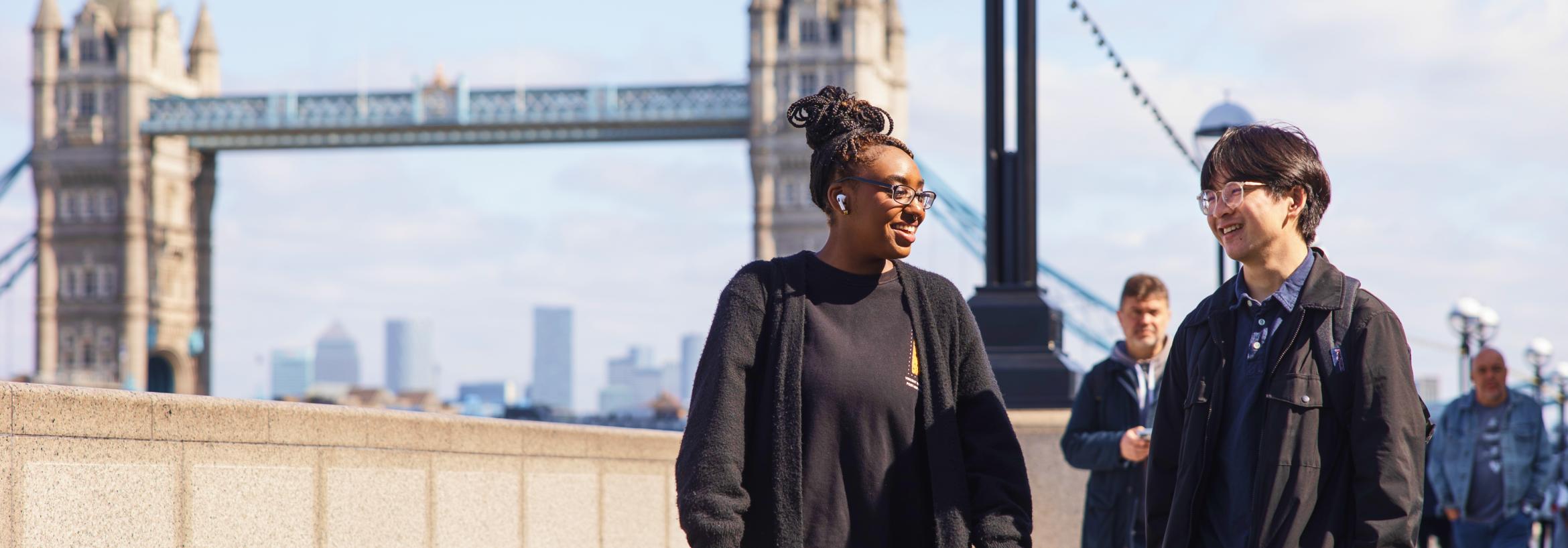 Two people walk and talk on a sunny day with Tower Bridge in the background; another person stands behind them on the sidewalk.