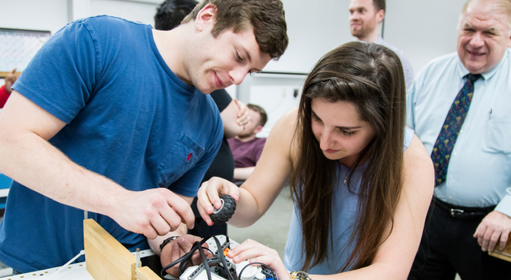 Two students working together on a hands-on project in a classroom while others observe in the background.