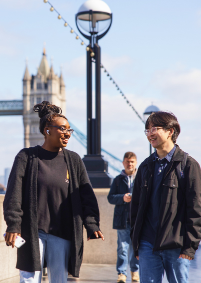 Two people walk and talk on a sunny day with Tower Bridge in the background; another person stands behind them on the sidewalk.